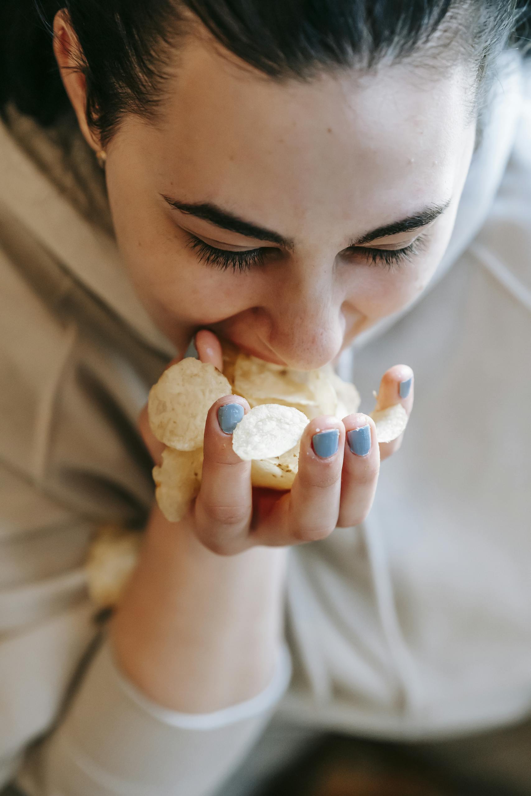 From above of crop female in sweater with black hair and handful of potato chips eating unhealthy fast food on blurred background